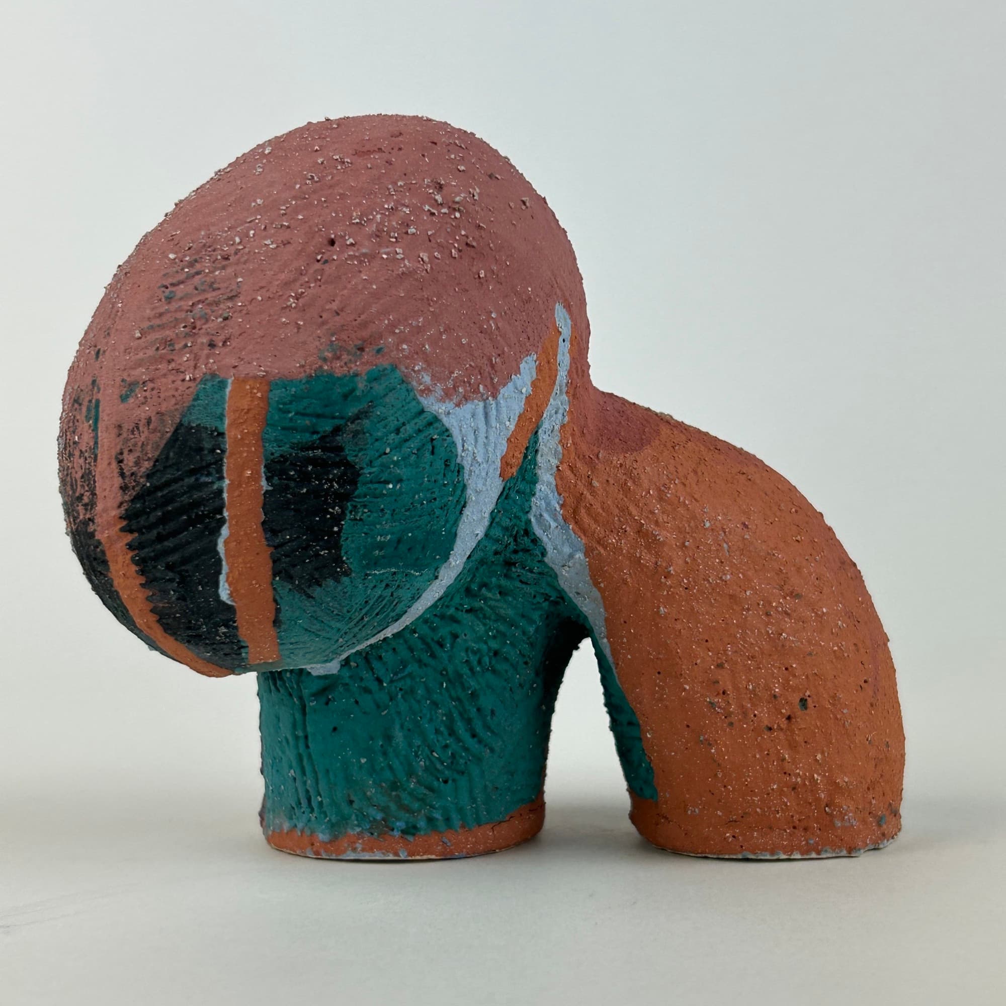Terracotta ceramic sculpture with a large bulbous head, washed in deep teal and pale blue over the raw red clay.