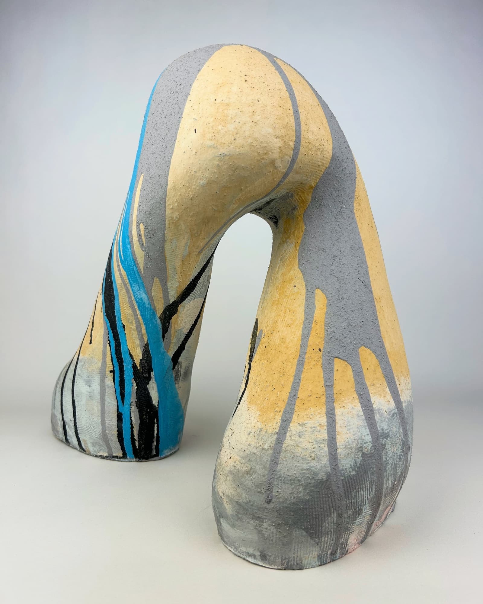 Tall arching ceramic sculpture in grey and pale yellow, with turquoise and black glaze streaking down one leg.