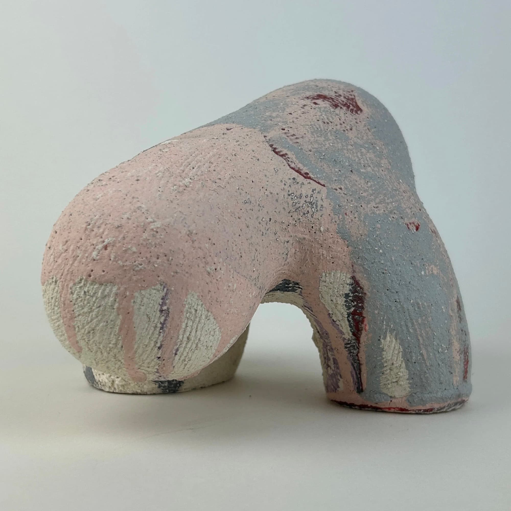 Hand-built ceramic sculpture: a low arching form in matte pink and grey with cream and oxblood glaze running down its flanks.