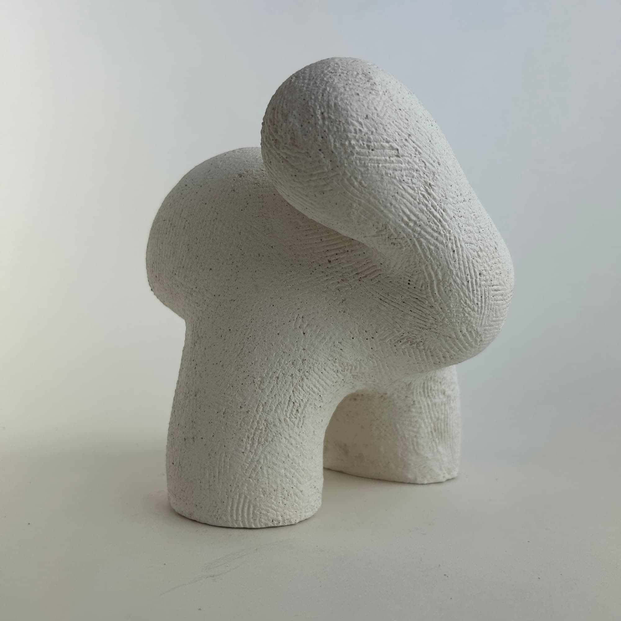 Chalk-white ceramic sculpture with a heavily textured surface, two rounded lobes meeting over an open arch.
