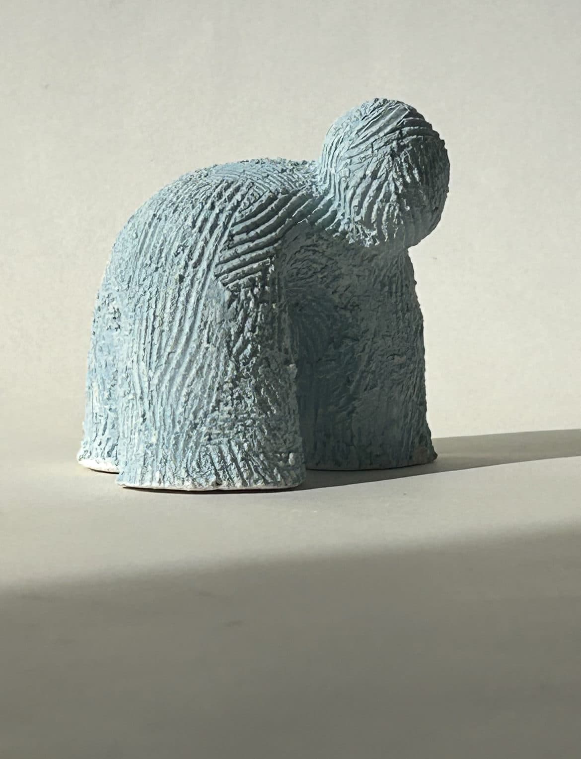 Pale blue ceramic sculpture with a heavily combed surface, the head folded over the body.